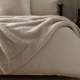 Olann Plain Velvet Throw in Natural, draped over a bed showing its shearling-look faux fleece front and smooth velvet reverse, with pillows and headboard in the background.