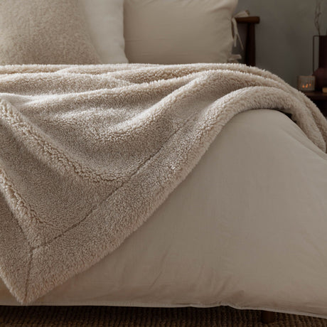 Olann Plain Velvet Throw in Natural, draped over a bed showing its shearling-look faux fleece front and smooth velvet reverse, with pillows and headboard in the background.