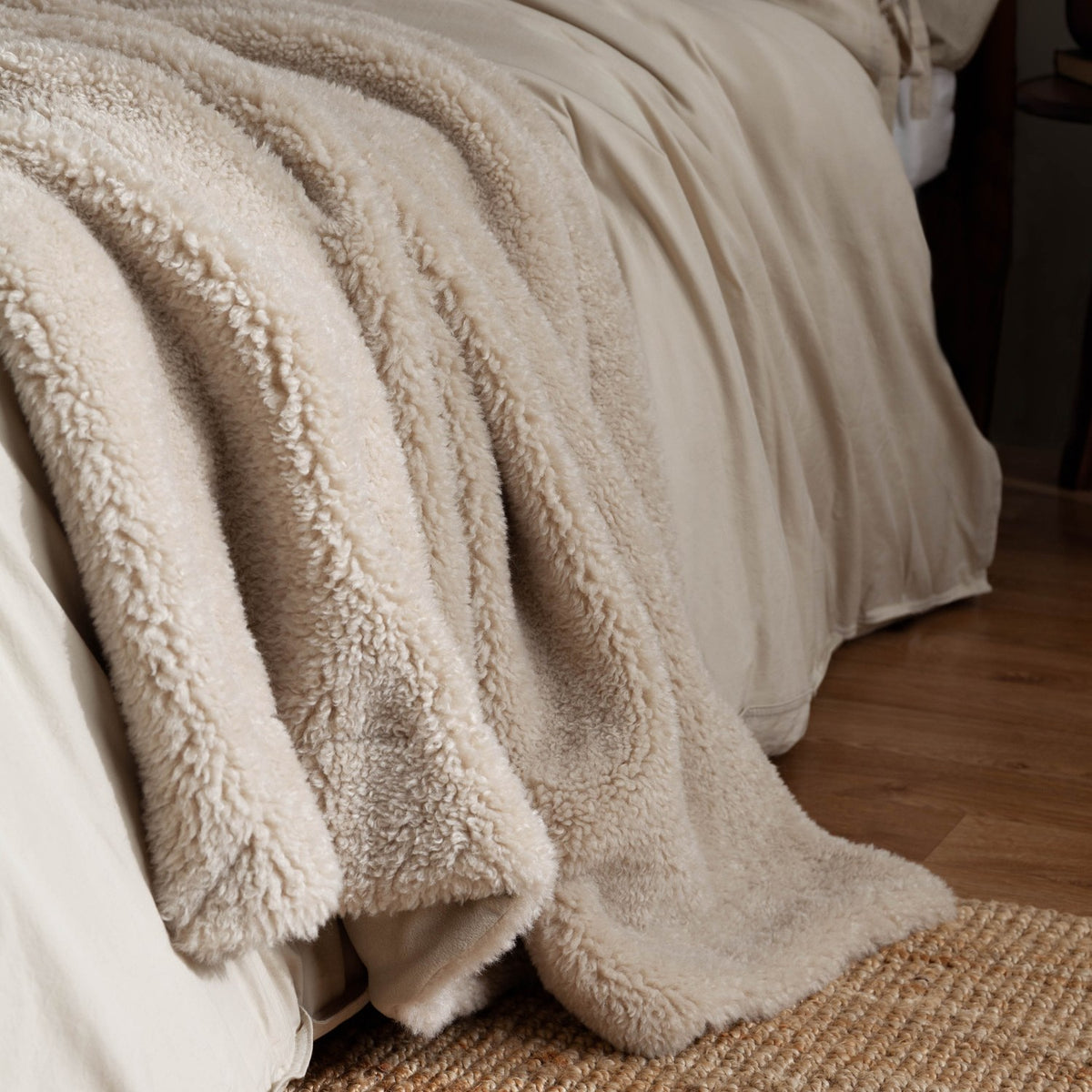 Olann Plain Velvet Throw in natural draped over a bed, showing the shearling-look faux fleece front against smooth linen bedding and a jute rug.