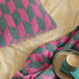 Olsen Geometric Polyester Cushion in Retro Pink shown on a bed with matching throw, both featuring a bold pink and teal geometric knit pattern, styled beside a striped yellow duvet.