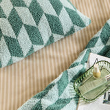 Olsen Geometric Polyester Throw in green tones with a tufted diamond pattern, edge visible on a beige striped bed sheet beside a magazine and green tray with beauty products.