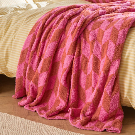 Olsen Geometric Polyester Throw in Pink Pop, a plush textured blanket with an interlocking chevron pattern in pink and rust, draped over a bed with striped yellow linen.
