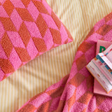Olsen Geometric Polyester Throw in Pink Pop, textured knit with interlocking pink and rust chevrons, matching cushion, styled on striped bedding with books.