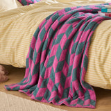Olsen Geometric Polyester Throw in Retro Pink and teal draped over a bed with striped yellow bedding, its tessellating diamond pattern creating a bold, textured knitted finish.
