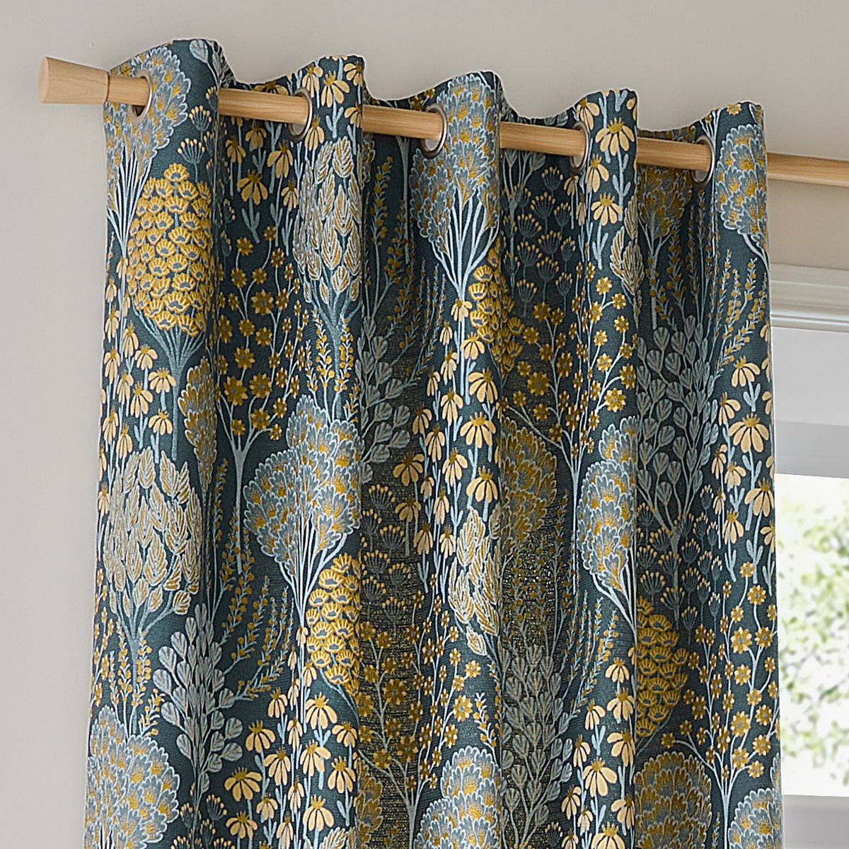 Ophelia Jacquard eyelet curtains in blue and saffron, hung on a wooden pole with metal grommets, showing a woven floral tree and daisy pattern near a window.