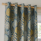 Ophelia Jacquard eyelet curtains in blue and saffron, hung on a wooden pole with metal grommets, showing a woven floral tree and daisy pattern near a window.