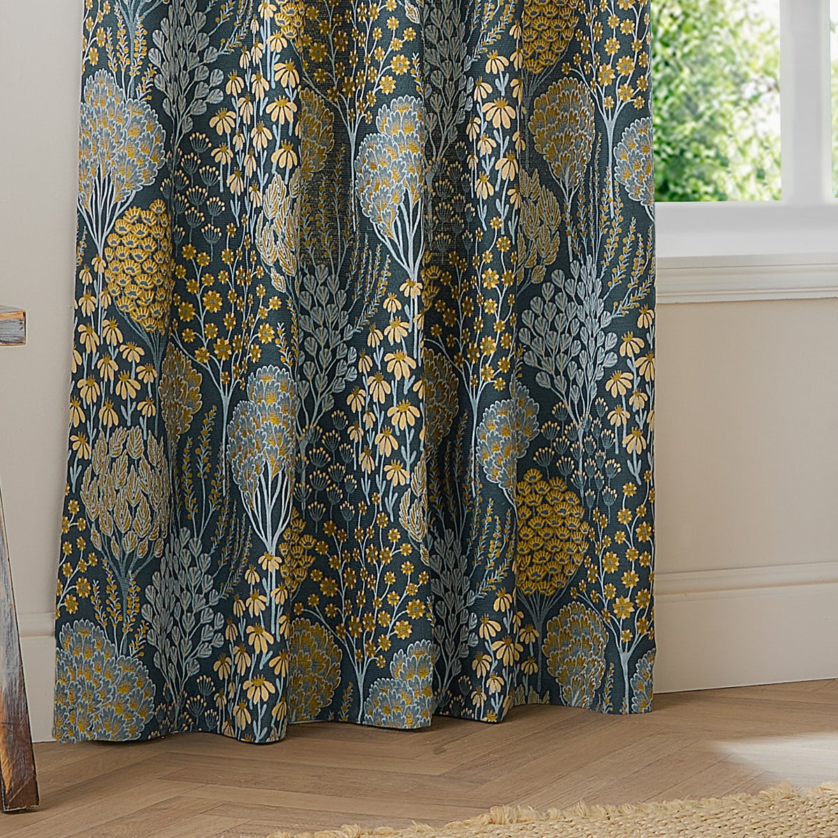 Ophelia Jacquard Fully Lined Eyelet Curtains in Blue/Saffron, shown gathered by a window with a woven floral tree and daisy pattern on a deep teal-blue ground.