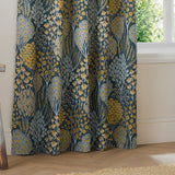 Ophelia Jacquard Fully Lined Eyelet Curtains in Blue/Saffron, shown gathered by a window with a woven floral tree and daisy pattern on a deep teal-blue ground.