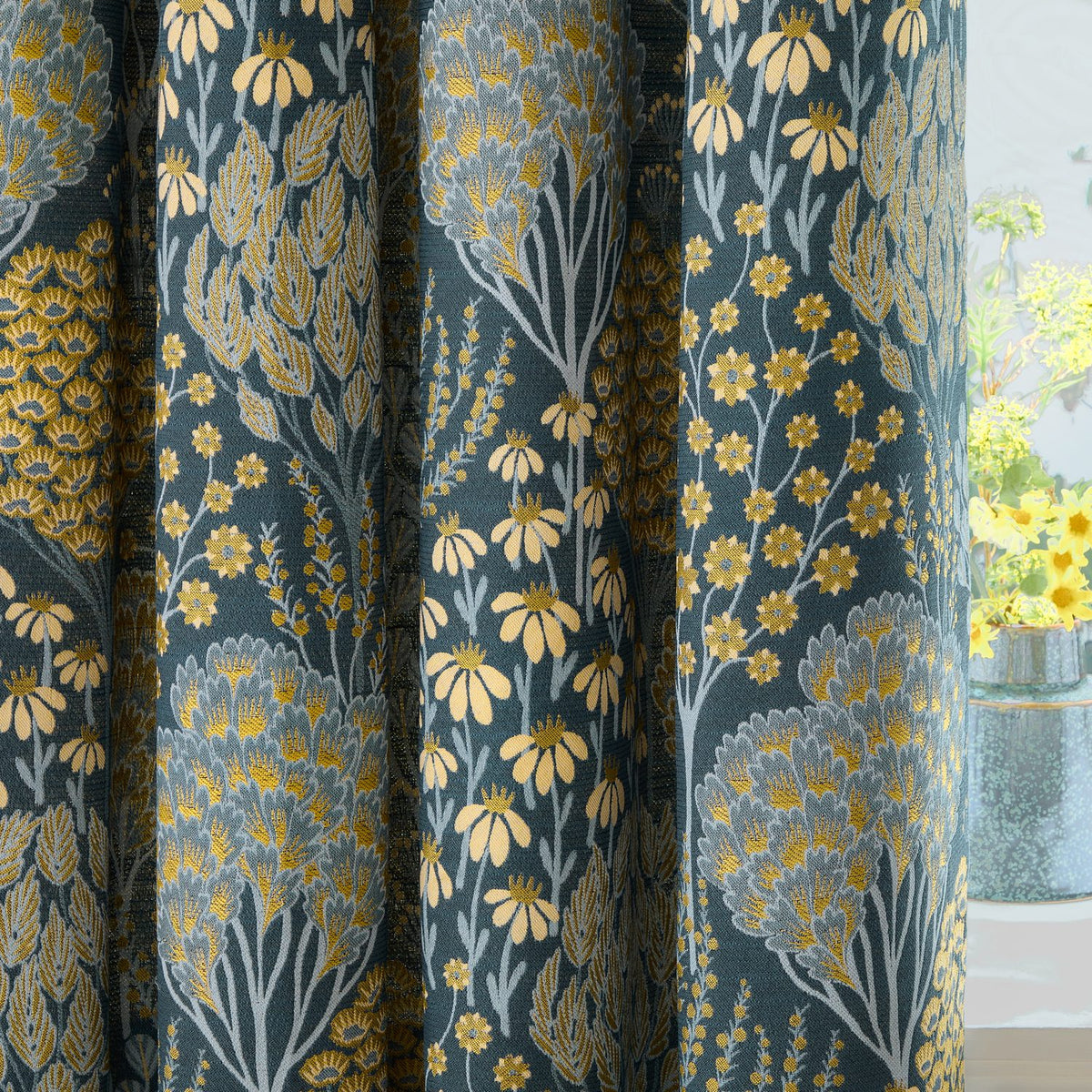 Ophelia Jacquard Fully Lined Eyelet Curtains in Blue/Saffron, shown draped in folds with a woven floral pattern of daisies and stylised trees in yellow, grey and blue on a teal ground.