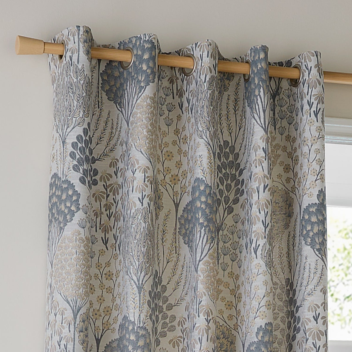 Ophelia Jacquard fully lined eyelet curtains in Wedgewood, showing a woven floral tree and daisy pattern in slate blue, gold and taupe on a pale ground, hung on a wooden pole.