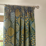 Ophelia Jacquard Polyester Pencil Pleat Curtains in blue and saffron, showing woven floral and tree motifs, hung pleated on a metal pole in a room setting.
