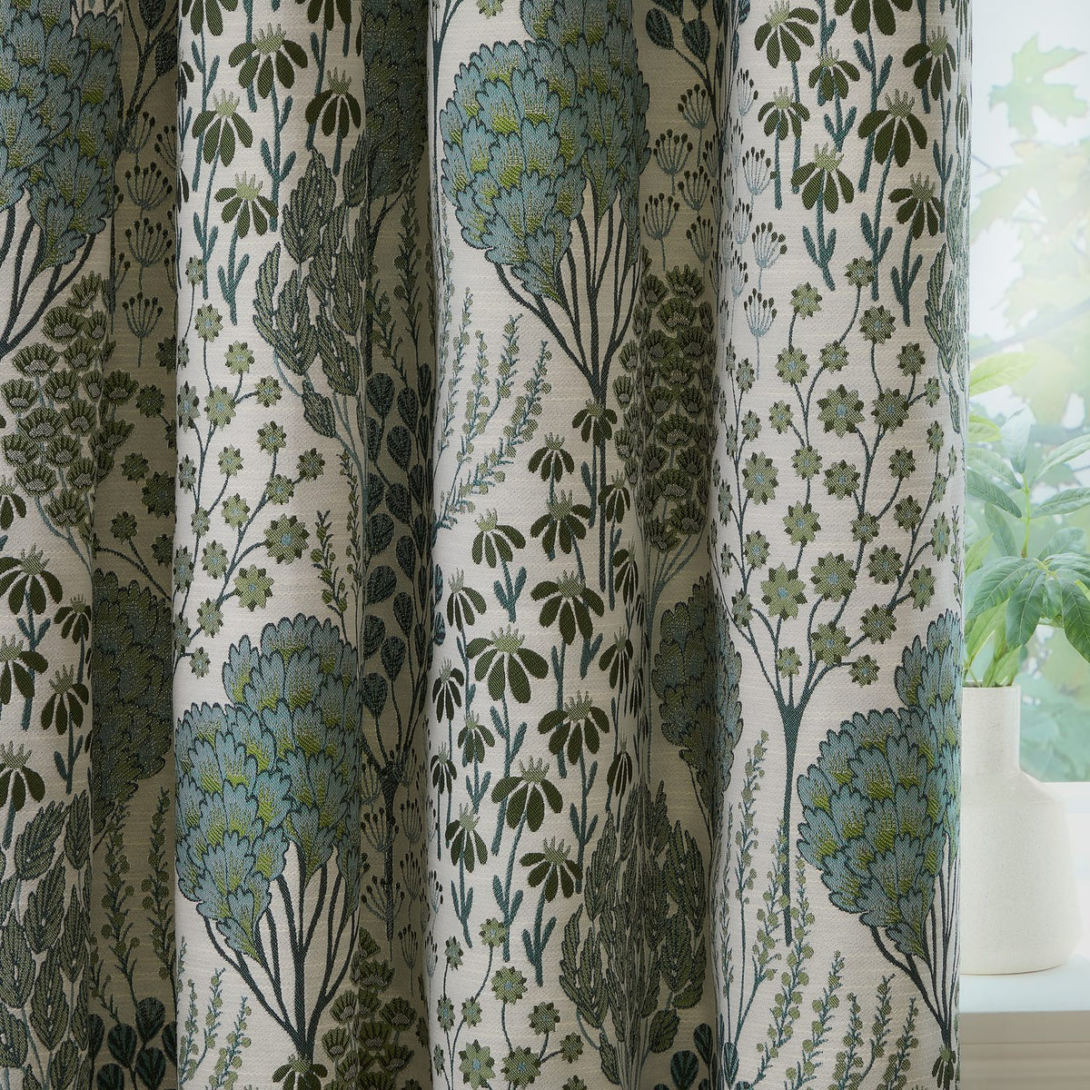 Ophelia Jacquard Pencil Pleat Curtains in Thyme/Mineral, featuring a woven botanical pattern of trees and wildflowers in green and blue-grey on cream fabric, styled by a window with a potted plant.