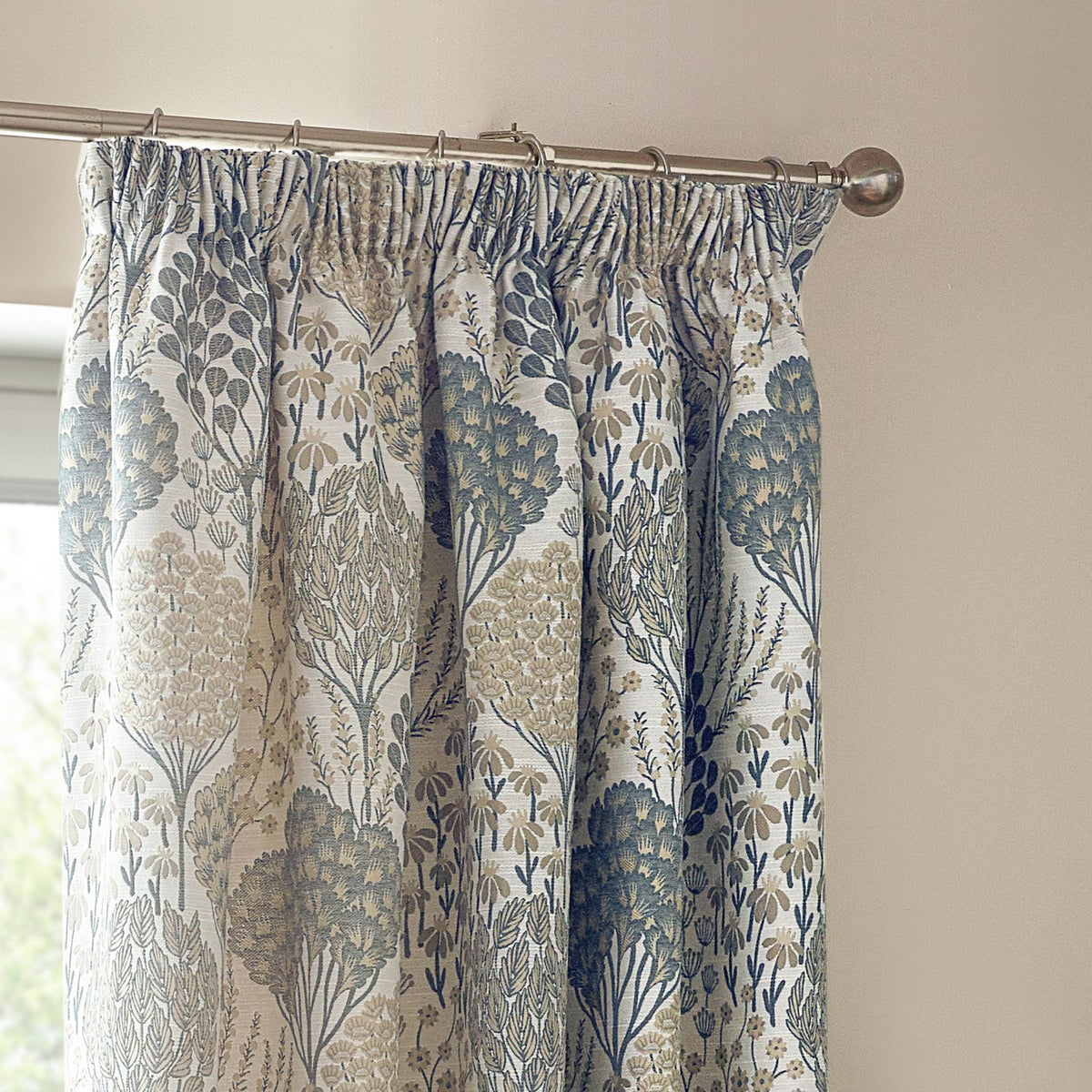 Ophelia Jacquard pencil pleat curtains in Wedgewood, showing a botanical tree and floral pattern in blue and beige on pale fabric, hung on a chrome curtain pole.