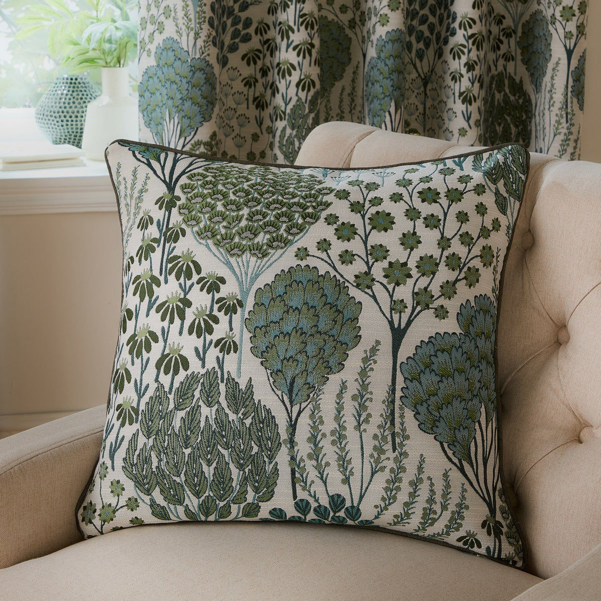 Ophelia Jacquard Polyester Cushion in Thyme/Mineral, square with cream ground and green and blue-teal tree, fern and seed head motifs, shown on a beige armchair.