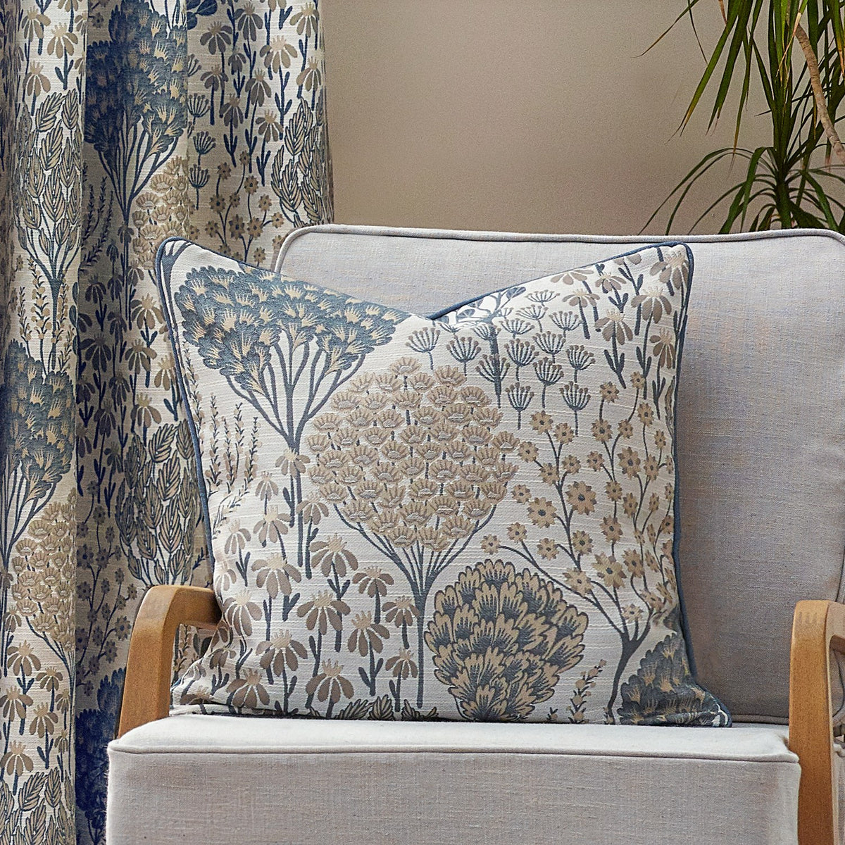 Ophelia Jacquard Polyester Cushion in Wedgewood, a square cushion with a woven blue and beige botanical pattern on off-white, piped trim, shown on a light armchair beside matching curtains.
