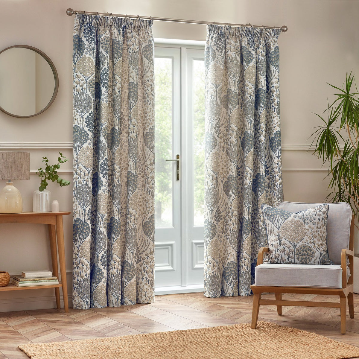 Ophelia Jacquard Polyester Cushion in Wedgewood on a light armchair, showing a woven botanical pattern in blue and beige with piped trim, in a living room with curtains and wood flooring.