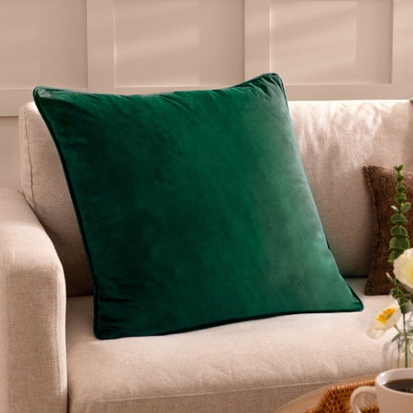Opulence plain velvet square cushion in bottle green with piped edge, shown on a beige sofa in a living room setting.