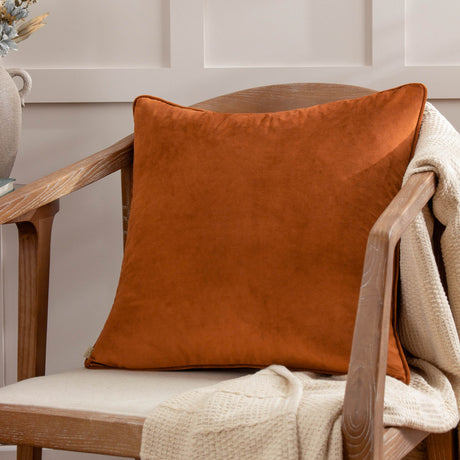 Opulence Plain Velvet Cushion in ginger orange with piped edges, square shape, styled on a wooden armchair with a cream knit throw.