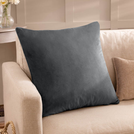 Square granite grey velvet cushion with piped edge, shown on a cream sofa in a living room setting beside a vase and side table.