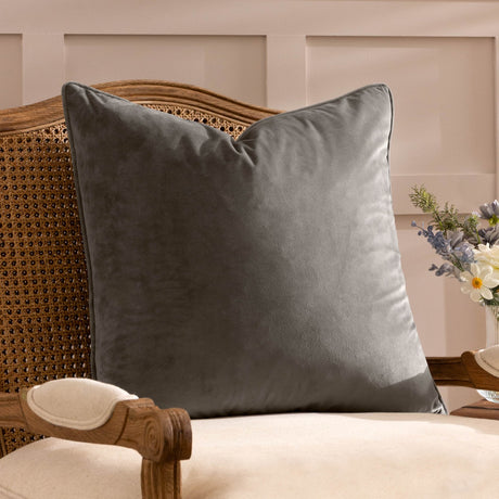 Opulence Plain Velvet Cushion in mink brown with piped edge, styled on a rattan chair beside a cream armrest and flowers.