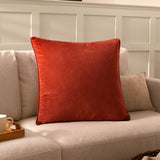 Opulence Plain Velvet Cushion in Sunset rust-orange with piped edge, styled on a neutral sofa in a living room setting.