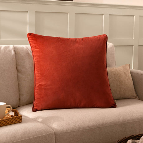 Opulence Plain Velvet Cushion in Sunset rust-orange with piped edge, styled on a neutral sofa in a living room setting.