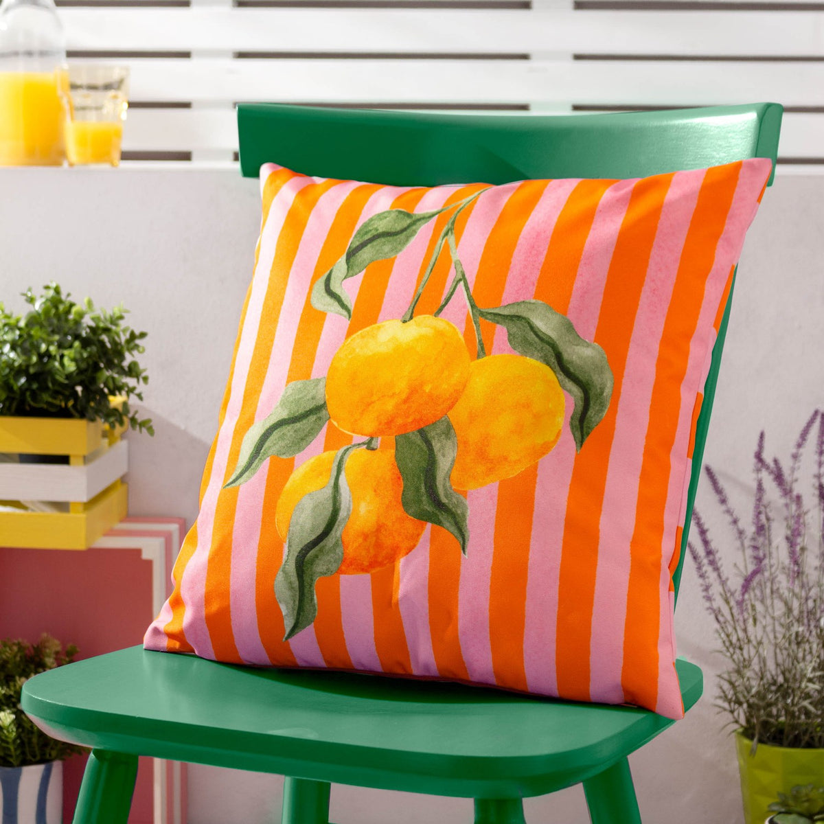 Square polyester outdoor cushion with pink and orange stripes and a watercolour orange branch design, shown on a green chair on a patio.