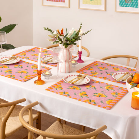 Rectangular pink placemats with an orange fruit print and striped border, set on a dining table with white plates, gold cutlery, candles, and floral centrepiece.