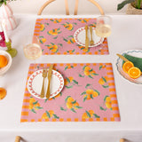 Two pink woven polyester placemats with an orange fruit and leaf print, bordered by orange and pink stripes, styled on a dining table with plates, cutlery and wine glasses.