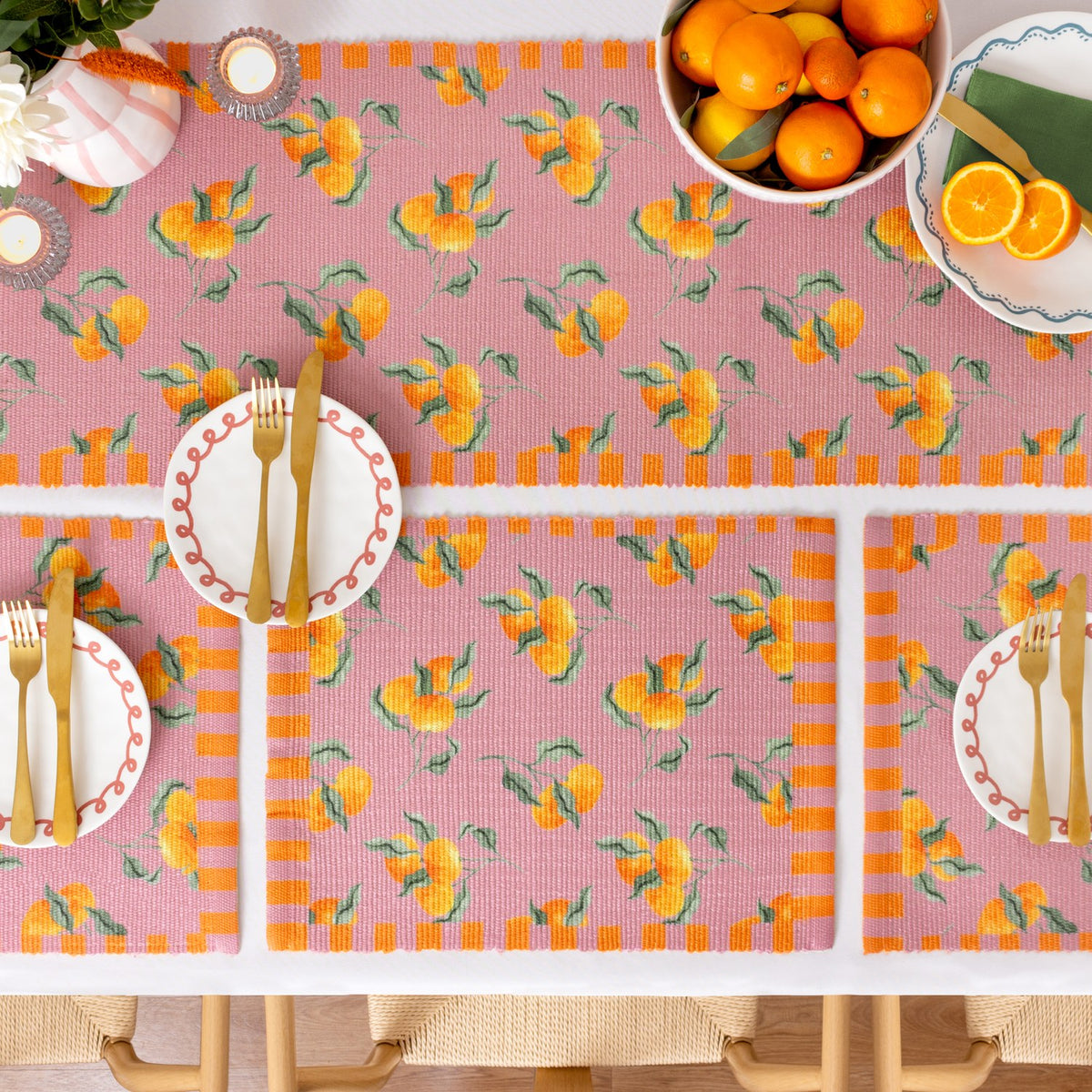 Rectangular pink polyester placemats with an orange fruit print and striped orange border, styled on a dining table with plates, cutlery, and a fruit bowl.