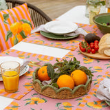 Oranges Floral Polyester Placemats in pink with striped border, styled on an outdoor dining table with a woven fruit basket, citrus-print cushion, and place settings.