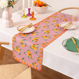 Pink polyester table runner with a scattered orange and green leaf print, bordered by orange striped ends, styled on a dining table with matching place settings.