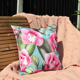 Orchids Floral Polyester Cushion in Duck Egg, square scatter cushion with pink orchid print and green leaves on blue polyester fabric, shown outdoors on a peach throw.