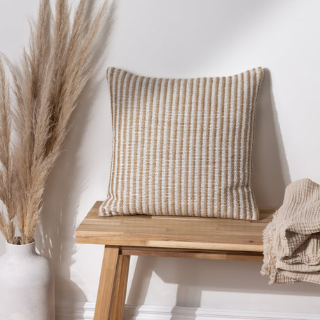 Organik Plain Cotton Cushion in Natural, a square jute and cotton cushion with fine woven stripes, shown on a wooden bench beside pampas grass in a light neutral room.