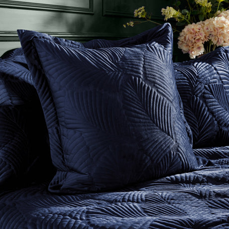 Palmeria Velvet Cotton Cushion in navy with a quilted, embroidered palm-leaf design, styled on a matching navy velvet bedspread beside pale flowers.
