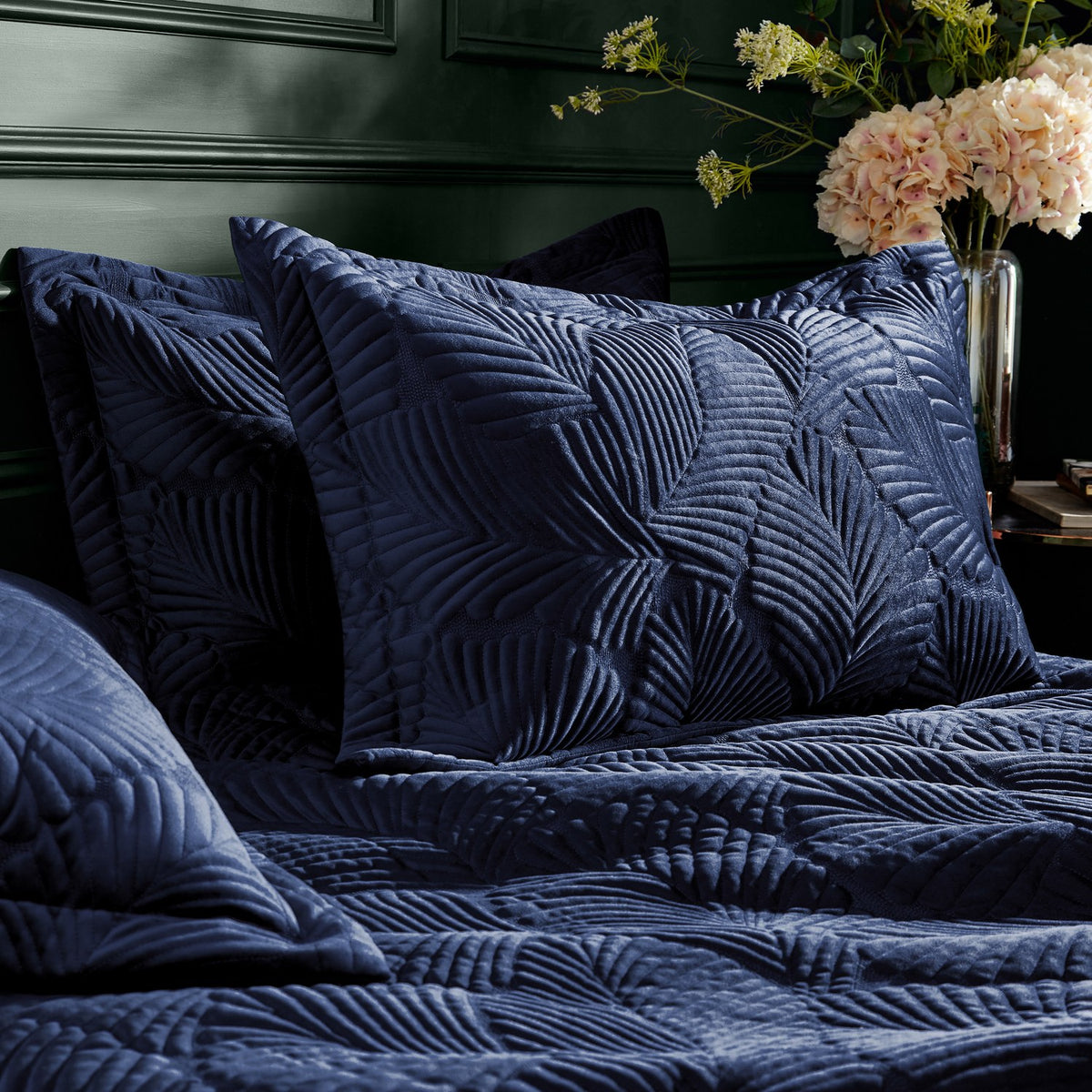 Palmeria Velvet Jungle Duvet Cover Set in navy on a bed, with matching quilted pillow shams showing raised leaf-pattern stitching, set in a bedroom with dark green panelled wall.