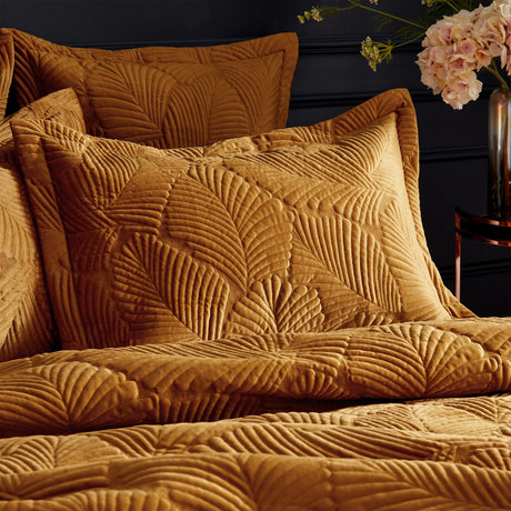 Palmeria Velvet Cotton Pillowcase Pair in gold, shown on a bed with quilted palm-leaf stitching and an oxford edge border, set against a dark background with soft florals nearby.