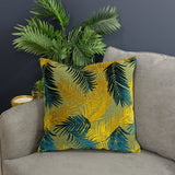 Palm Grove Jacquard Velvet Cushion in gold and teal with a woven palm leaf pattern, shown on a grey sofa beside a potted palm plant.