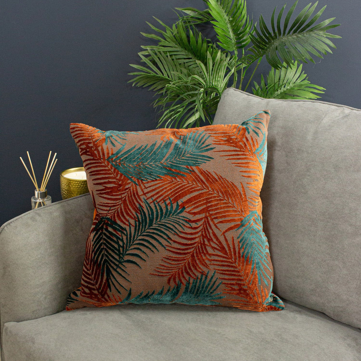 Palm Grove Jacquard Velvet Cushion in teal and rust palm leaf pattern on a mid-brown base, shown on a grey sofa beside a reed diffuser and green plant.