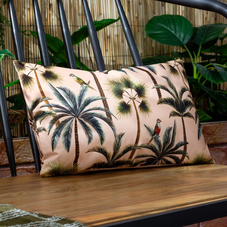Rectangular blush pink outdoor cushion with a tropical palm tree and parrot print, styled on a wooden bench against a black spindle-back chair.