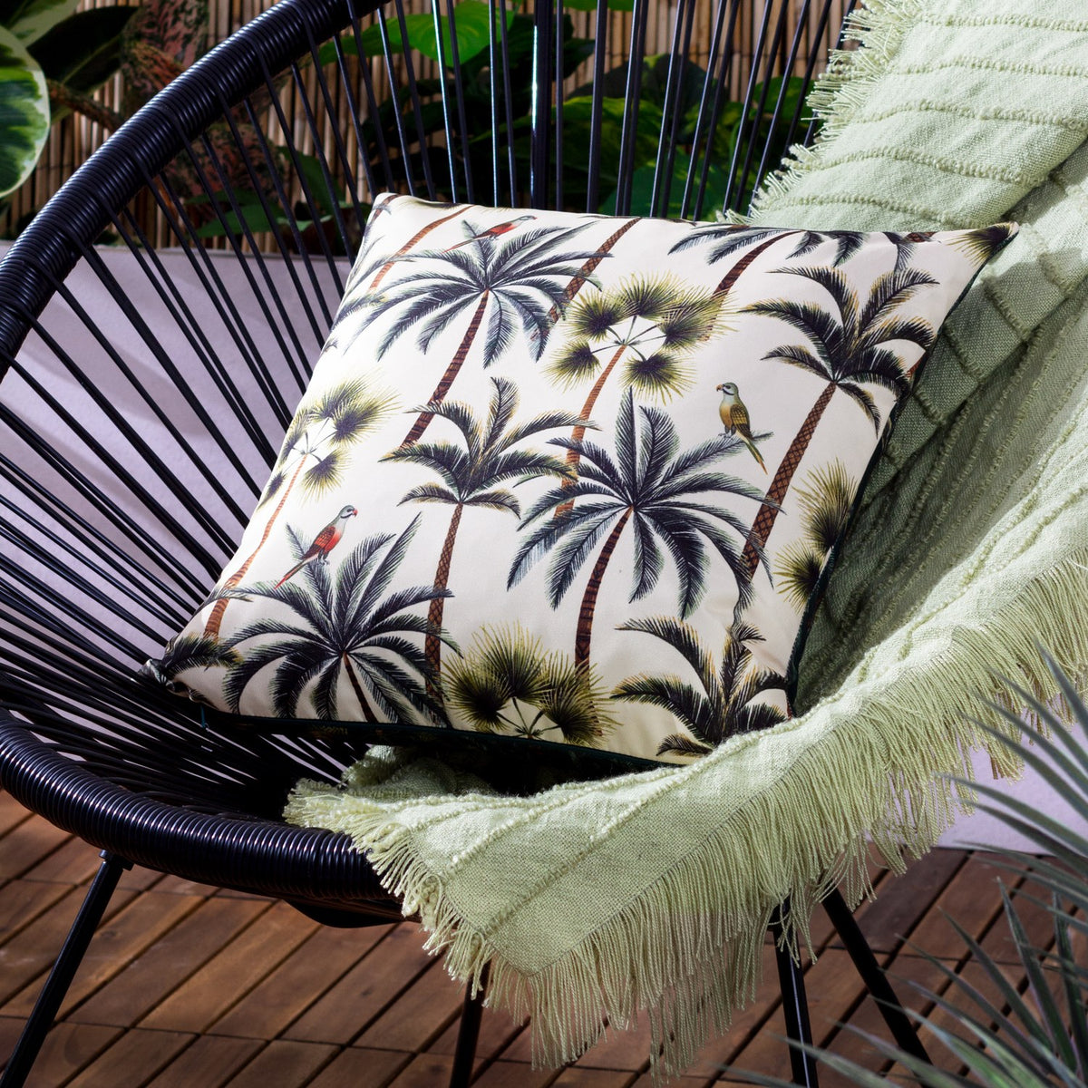 Palms Reversible Polyester Cushion in Forest, a square outdoor scatter cushion with cream and green tropical palm and parrot print, shown on a black woven chair with a light green fringed throw.