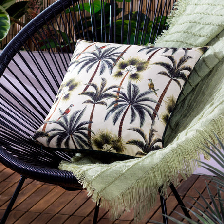 Palms Reversible Polyester Cushion in Forest, a square outdoor scatter cushion with cream and green tropical palm and parrot print, shown on a black woven chair with a light green fringed throw.