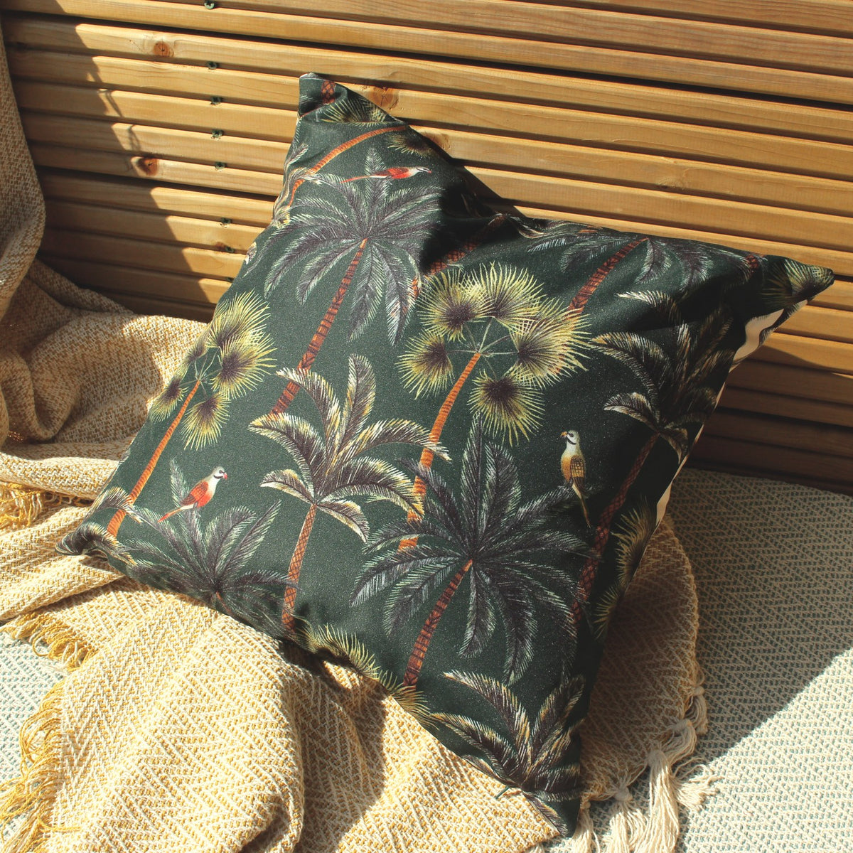 Palms Reversible Polyester Cushion in Forest green, showing a tropical palm and parrot print, styled against a slatted wooden bench with a woven throw.