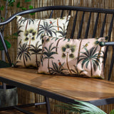 Two rectangular Palms cushions with tropical palm and parrot print in cream and blush pink tones, shown on a black metal bench against a bamboo backdrop.