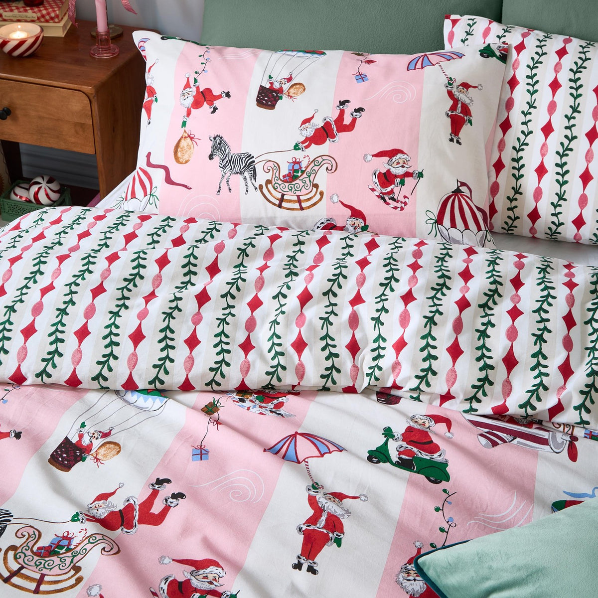 Passport To The North Pole duvet cover set in Candy Floss/Linen with pink Santa-print pillowcases and duvet reversing to a red mistletoe stripe, styled on a bed with a green headboard.