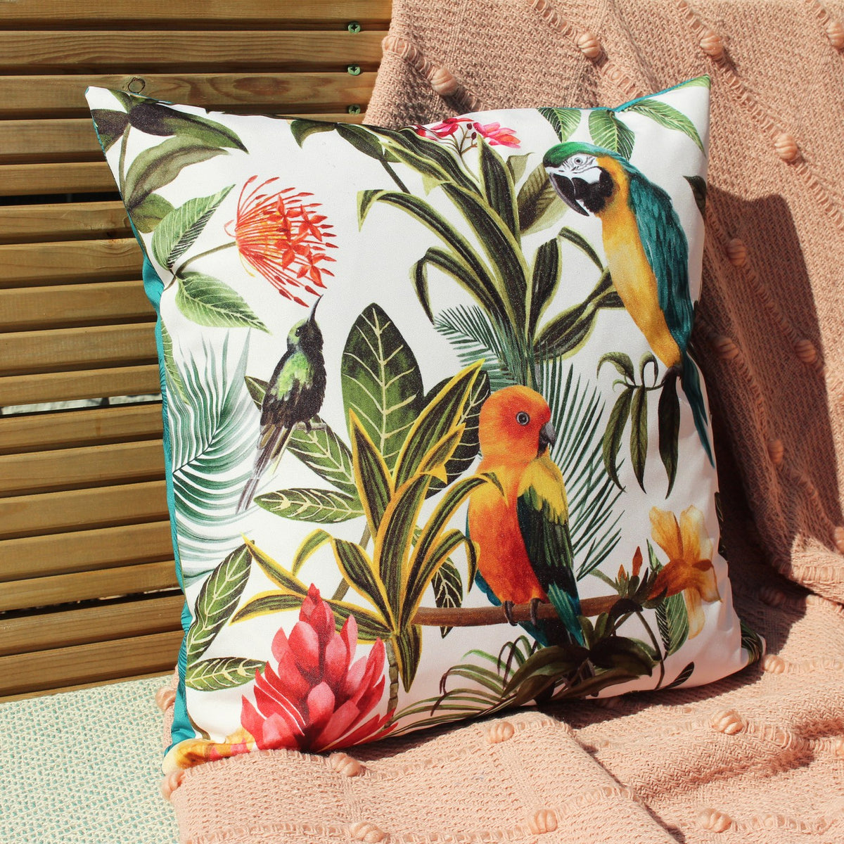 Square polyester cushion in white, teal-piped, printed with tropical parrots, foliage and flowers, shown outdoors against wooden slats and a peach throw.