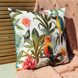 Square polyester cushion in white, teal-piped, printed with tropical parrots, foliage and flowers, shown outdoors against wooden slats and a peach throw.