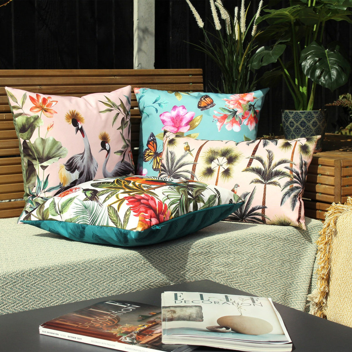 Parrots Animal Polyester Cushion in Multi/Teal with tropical parrot and foliage print on white, shown with patterned pillows on an outdoor bench.