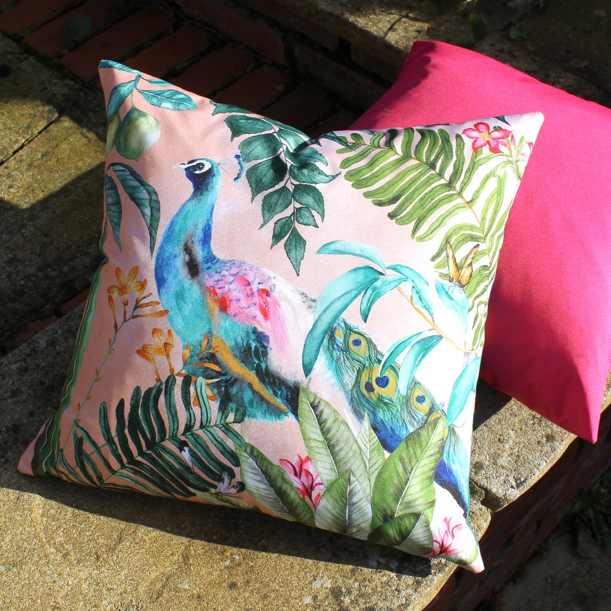 Square polyester outdoor cushion with a painted peacock among tropical leaves on blush pink, shown outdoors beside a pink cushion on stone.