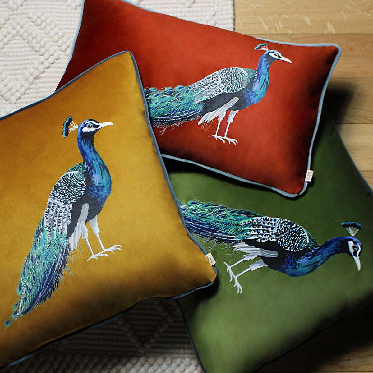 Square olive green velvet cushion with a printed peacock design, shown alongside mustard and red peacock cushions in matching style.